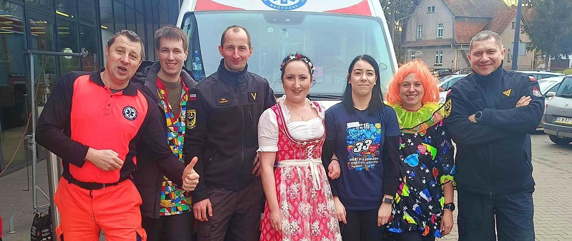 Strażacy i wolontariusze WOŚP w kolorowych strojach pozują do wspólnego zdjęcia przed karetką podczas finału Wielkiej Orkiestry Świątecznej Pomocy.