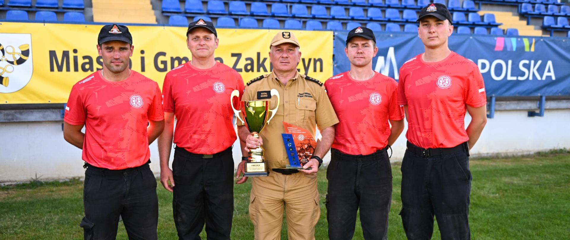 Na płycie boiska stoją strażacy ubrani w czarne spodnie i czerwone podkoszulki na głowach mają czapeczki. W środku komendant w piaskowym mundurze w rekach trzyma dyplom i puchar