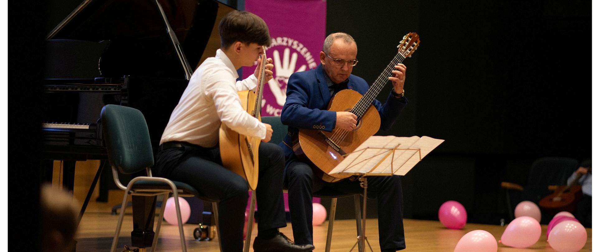 Kolorowe zdjęcia dwóch, siedzących osób i grających na gitarach klasycznych. Panowie znajdują się na scenie wśród balonów w różnych odcieniach różu. Obaj patrzą na nuty znajdujące się przed nimi na pulpicie.
