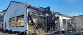 Zdjęcie przedstawia budynek - część warsztatowo-magazynową - po pożarze wewnątrz obiektu. Na zdjęciu widoczny również po lewej stronie samochód pożarniczy.