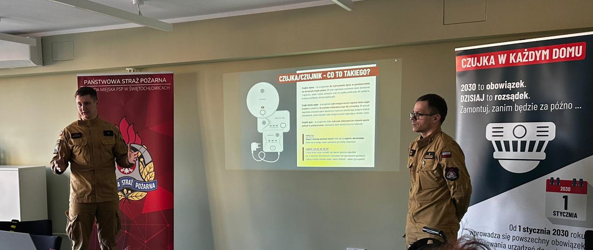 Zdjęcie przedstawia prelekcję prowadzoną przez dwóch strażaków z Państwowej Straży Pożarnej dla grupy seniorów. Funkcjonariusze w piaskowych mundurach stoją przy ścianie z wyświetloną prezentacją pt. „CZUJKA/CZUJNIK – CO TO TAKIEGO?”, omawiając rodzaje urządzeń zabezpieczających przed dymem i gazem. Obok nich znajduje się duży baner kampanii „Czujka w każdym domu”, który informuje o nadchodzącym obowiązku posiadania takich urządzeń od 2030 roku.
Uczestnicy spotkania siedzą tyłem do obiektywu przy białym stole konferencyjnym, uważnie słuchając wykładu. Na stole widoczne są materiały szkoleniowe, butelki z wodą oraz rzutnik, a całe pomieszczenie jest dobrze doświetlone światłem dziennym wpadającym przez duże okno. Spotkanie ma charakter edukacyjny i ma na celu zwiększenie świadomości o zagrożeniach związanych z pożarami oraz zatruciem tlenkiem węgla.

