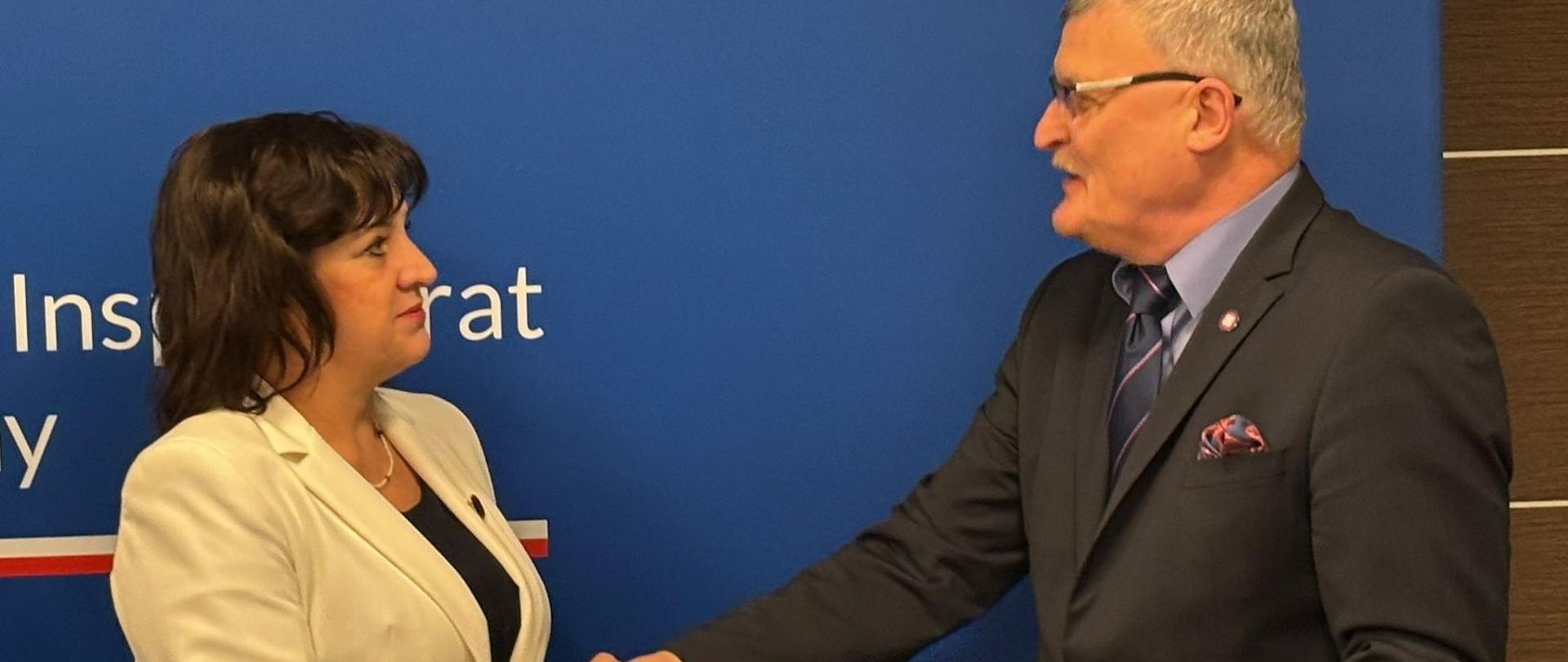 Na drugim planie niebieskie tło z orłem i napisem Główny Inspektorat Sanitarny oraz logo Państwowej Inspekcji Sanitarnej. Na jego tle dr n. med. Paweł Grzesiowski Główny Inspektor Sanitarny wręcza nominację na Zastępcę Podlaskiego Państwowego Wojewódzkiego Inspektora Sanitarnego w Białymstoku Pani dr Izabeli Katarzynie Szamrej. Oboje ściskają sobie dłonie. 