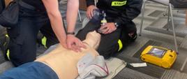 Na zdjęciu widzimy jak strażacy wykonują symulację resuscytacji krążeniowo- oddechowej u osoby dorosłej. Jeden z ratowników wykonuje 30 uciśnięć klatki piersiowej, natomiast drugi wykonuje 2 oddechy ratownicze za pomocą worka samorozprężalnego.