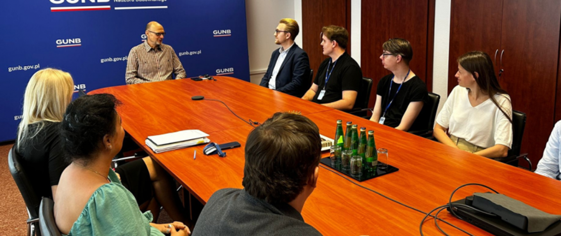 Grupa osób przy stole konferencyjnym podczas spotkania w GUNB, z widocznym banerem instytucji i materiałami biurowymi.