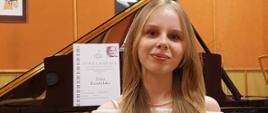 Zdjęcie kolorowe. Uśmiechnięta dziewczyna o blond włosach, ubrana w jasny sweter siedzi przed otwartym czarnym fortepianem. Na pulpicie fortepianu stoi dyplom laureata konkursu pianistycznego.