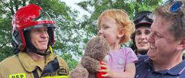 Na tarasie widokowym stoi tata trzymający na rękach córeczkę, która była uwięziona w zjeżdżalni. Dziewczynka ma w rękach maskotkę - misia, którego otrzymała od strażaków. Obok nich stoja strażacy w ubraniach specjalnych w kolorze piaskowym i czerwonych hełmach. W tle - drzewa.
