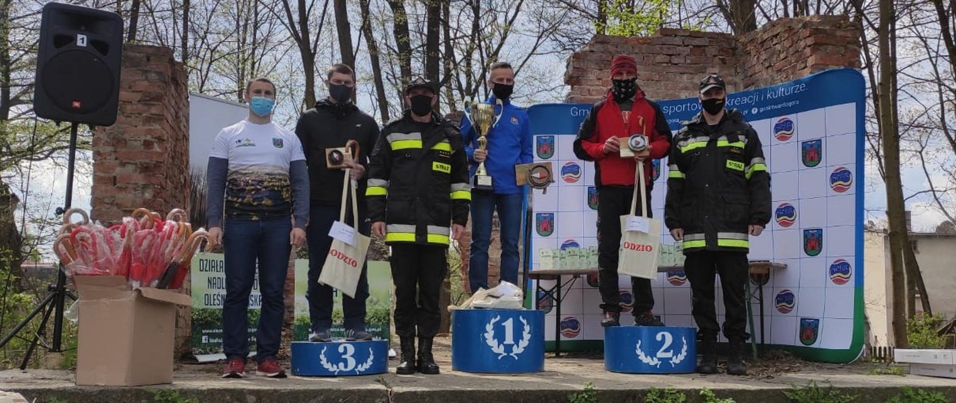 Na zdjęciu widać strażaków, zwycięzców biegu na orientację. Stoją na podium z otrzymanymi pucharami. 