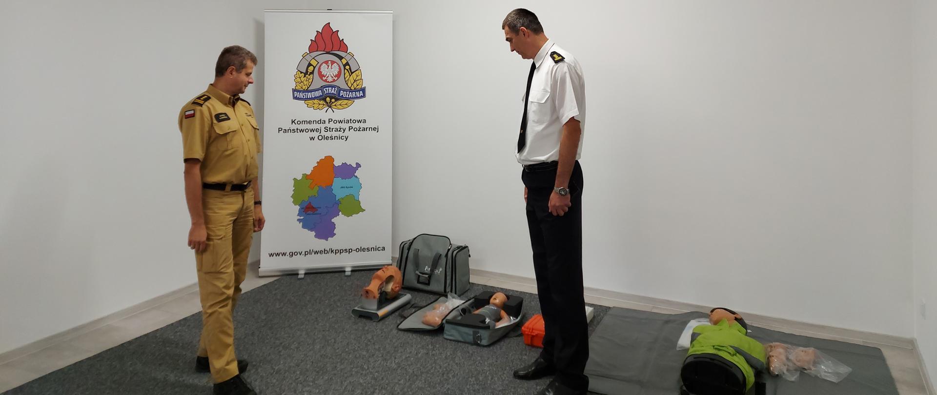 Na zdjęciu widać Dolnośląskiego Komendanta Wojewódzkiego PSP we Wrocławiu w mundurze piaskowym oraz Komendanta Powiatowego PSP w Oleśnicy w mundurze wyjściowym (biała koszula, czarne spodnie). Komendanci oglądają salę edukacyjną. 