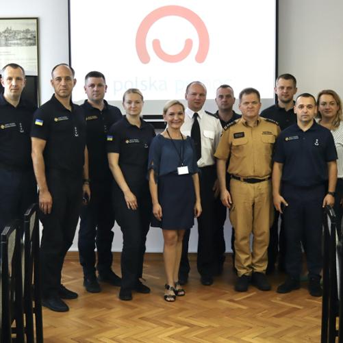 Zdjęcie uczestników projektu na tle loga Polska Pomoc, po bokach krzesła