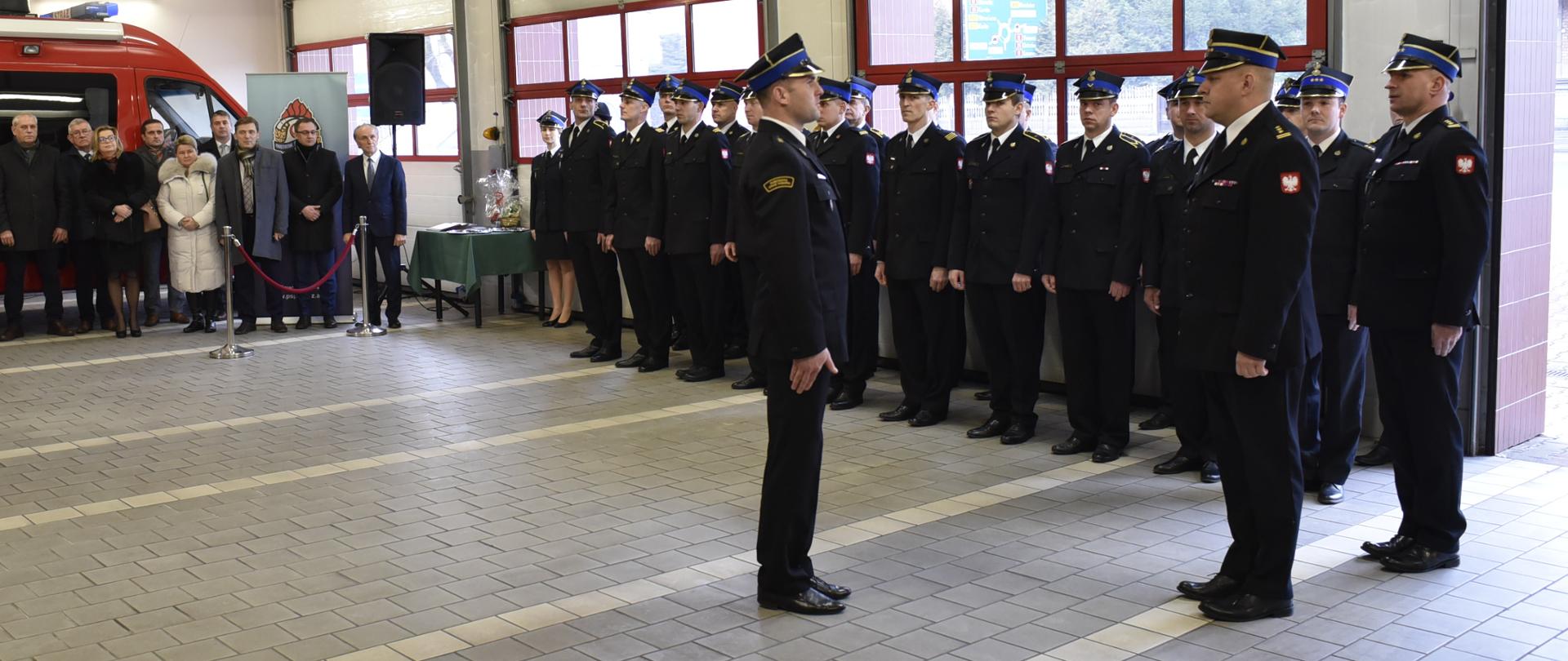 w garażu - składanie meldunku strażakowi przez innego strażaka, strażcy ubrani w mundury wyjściowe, i w rogatywkach na głowie za nimi stoją w szeregu stoją strażacy w mundurach wyjściowych i w rogatywkach, z prawej strony grupa stoi ludzi, za mimi czerwony samochód