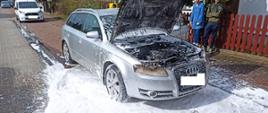 Centralny punkt zdjęcia stanowi auto osobowe. Auto srebrne, nadwozie kombi. Auto ma podniesioną maskę. Pożarem objęta była komora silnika. Dokoła auta piana.