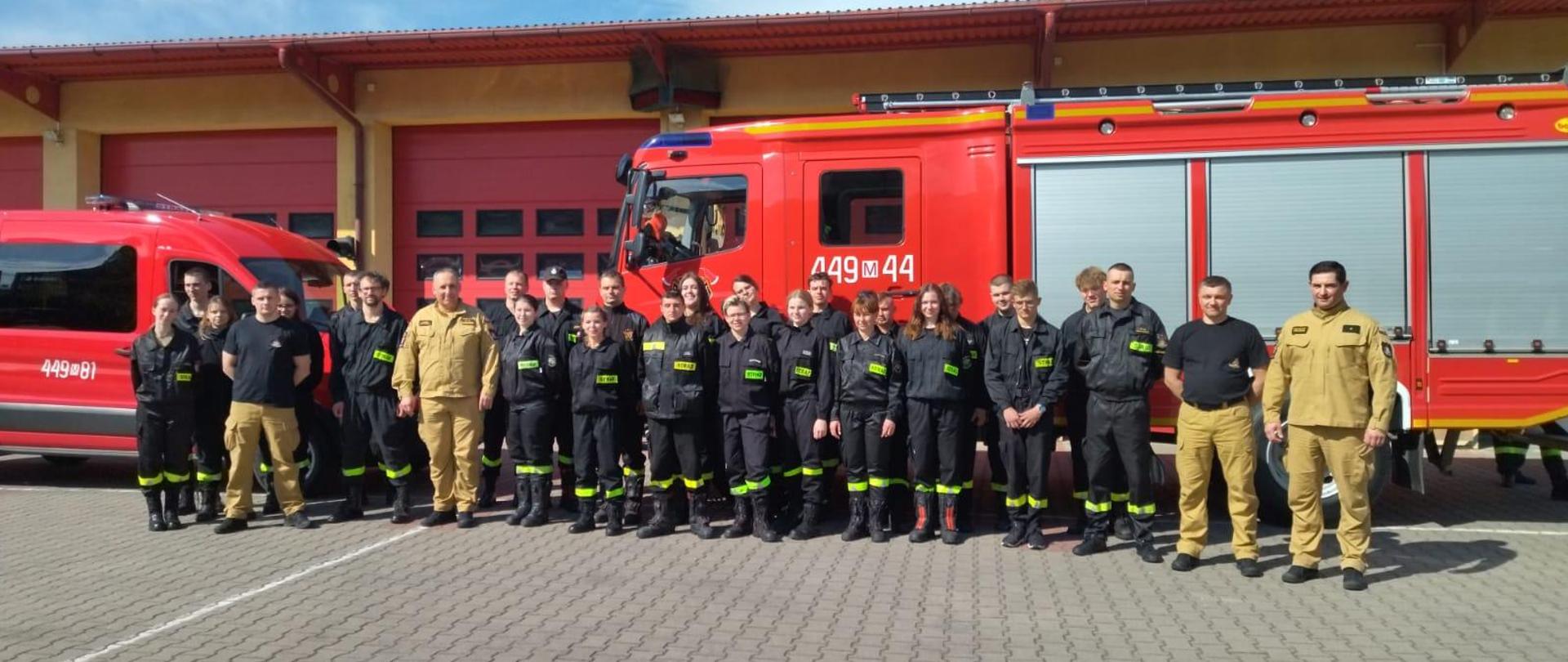 Uczestnicy szkolenia po zakończonych egzaminach stoją w dwuszeregu na tle s=dwóch samochodów pożarniczych.