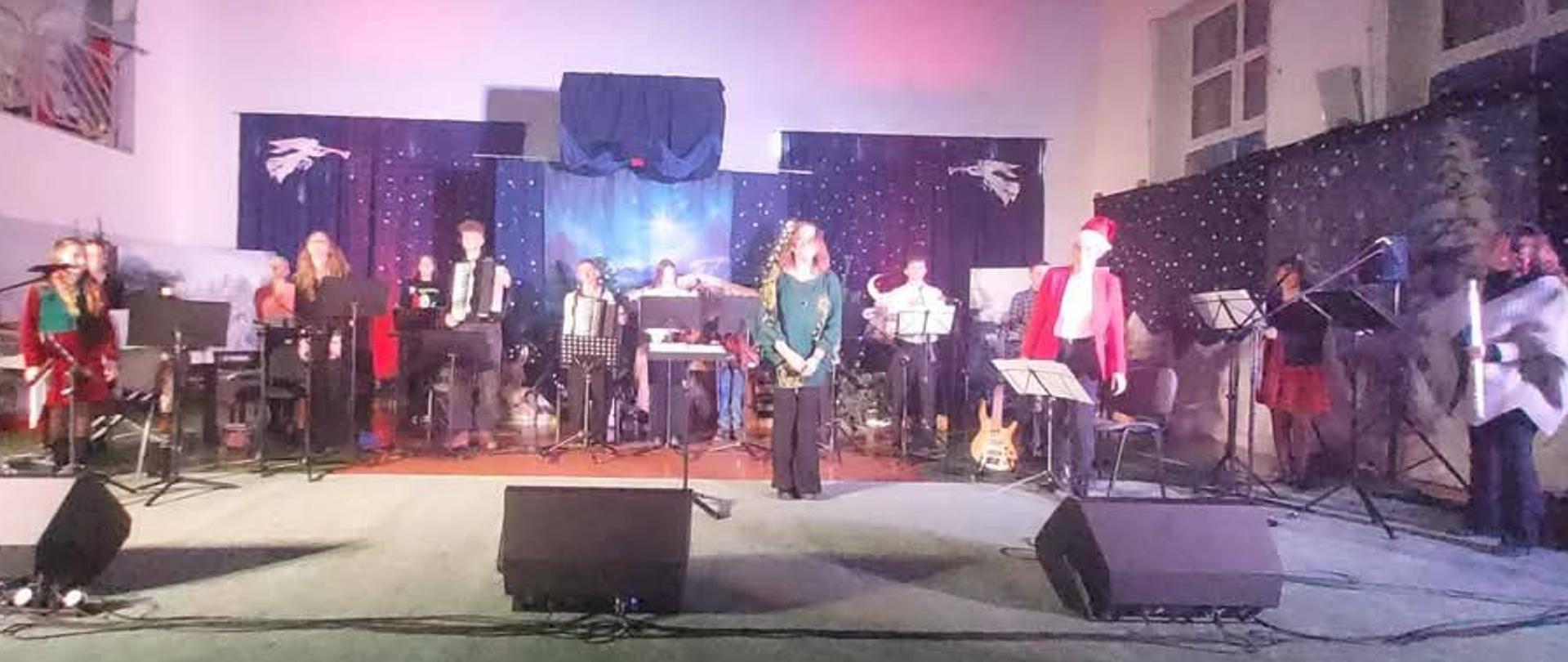 Zespół w całej okazałości na scenie. Wszyscy muzycy stoją z instrumentami, przed zespołem pani dyrygent.