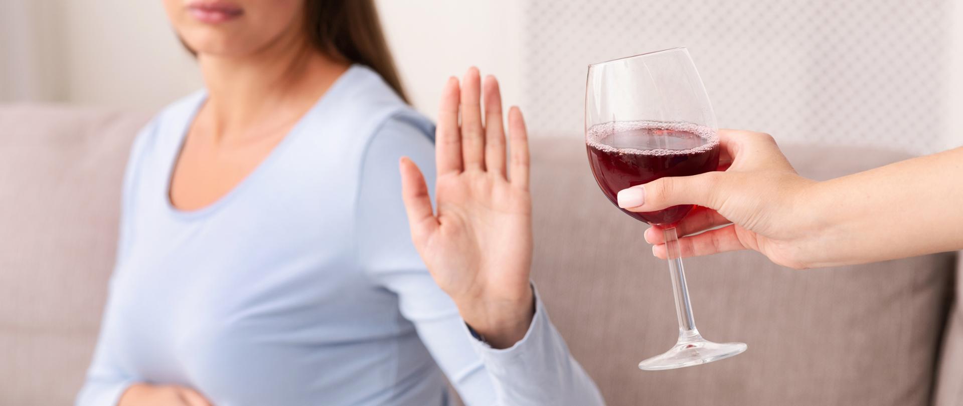Kobieta w ciąży, siedząca na kanapie w pomieszczeniu gestykulując, odmawia przyjęcia kieliszka z czerwoną substancją przypominającą wino. 