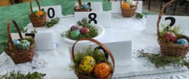 Szerokie ujęcie stołu z wystawą pisanek w koszyczkach. Obok każdego koszyczka stoi biała kartka z czarnym numerem identyfikacyjnym (m.in. 10, 8, 5, 2).