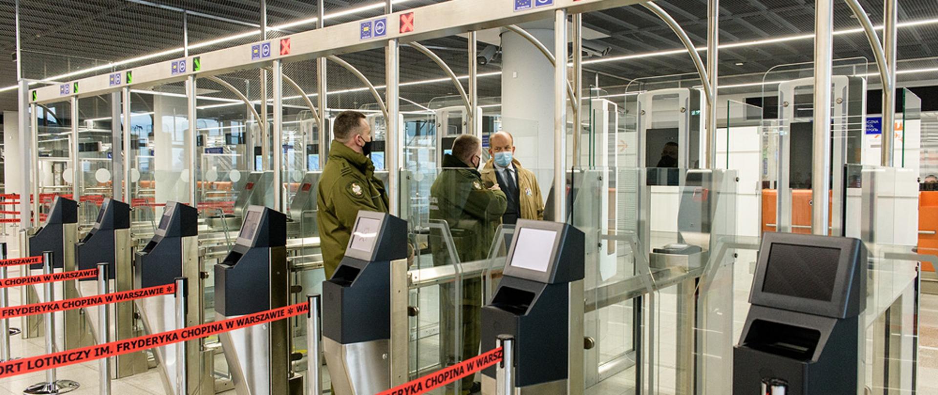 Zdjęcie przedstawia fragment spotkania, w trakcie którego przekazano kolejnych dziesięć bramek ABC, służących do automatycznej odprawy granicznej pasażerów Lotniska Chopina w Warszawie. Na pierwszym planie znajduje się linia bramek. Tuż przy niej znajduje się trzech mężczyzn - jeden ubrany w beżowy płaszcz i dwóch ubranych w ciemnozielone mundury Straży Granicznej.