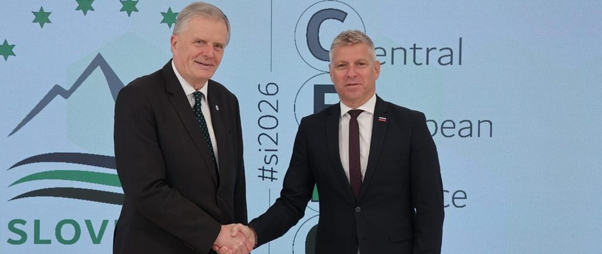 Dwaj przedstawiciele delegacji w garniturach podają sobie dłonie przed ścianką z oznaczeniami CEDC i Slovenia 2026.