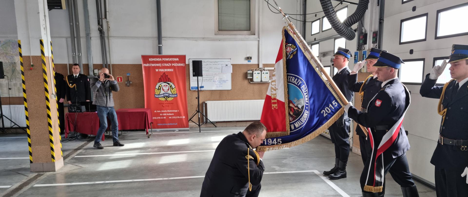 Strażak klęczy przed sztandarem Komendy Powiatowej Państwowej Straży Pożarnej w Malborku całując go. Sztandar trzyma funkcjonariusz, obok stoją strażacy.