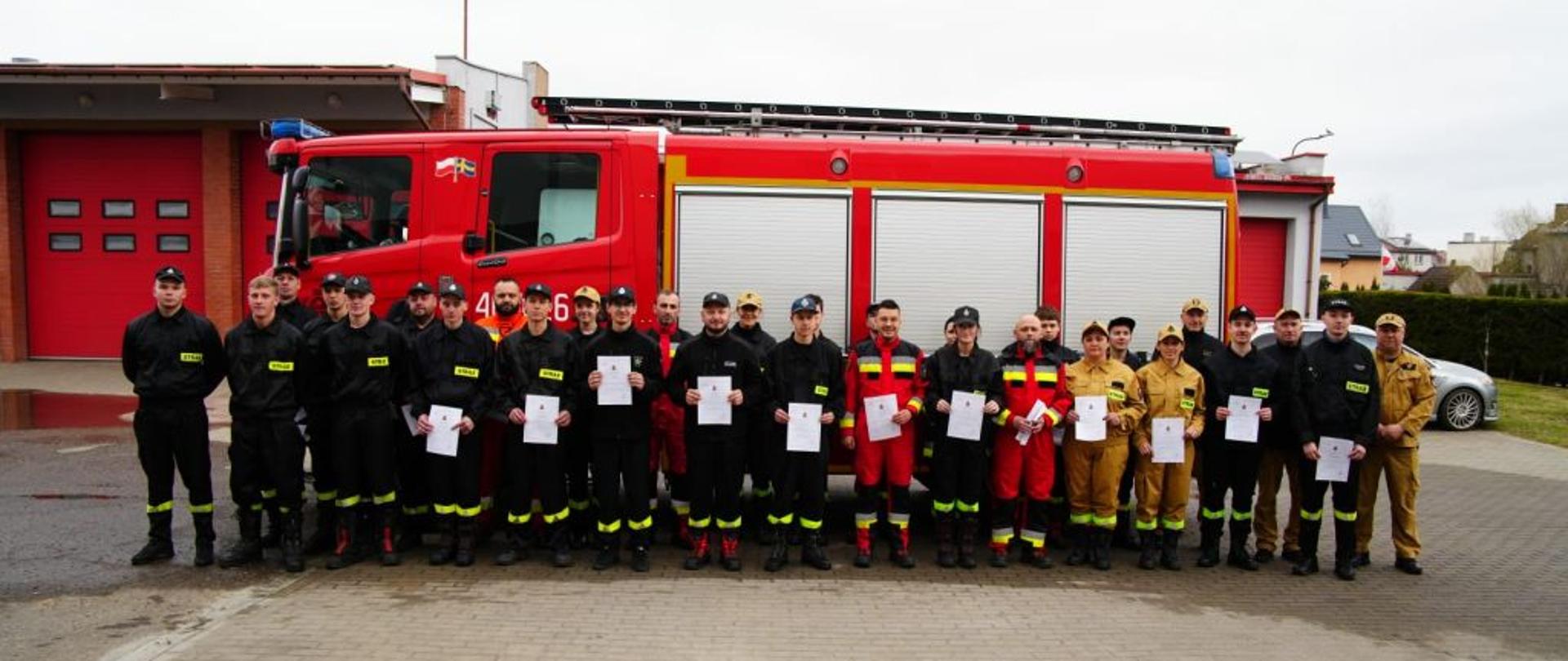Oficjalne zdjęcie grupowe uczestników szkolenia na placu manewrowym. Strażacy stoją w jednym rzędzie na tle dużego, czerwonego wozu gaśniczego marki Scania. Większość osób trzyma w dłoniach białe dyplomy. Po prawej stronie grupy stoją instruktorzy w beżowych mundurach. W tle widoczna jest bryła budynku straży pożarnej z czerwonymi bramami garażowymi.