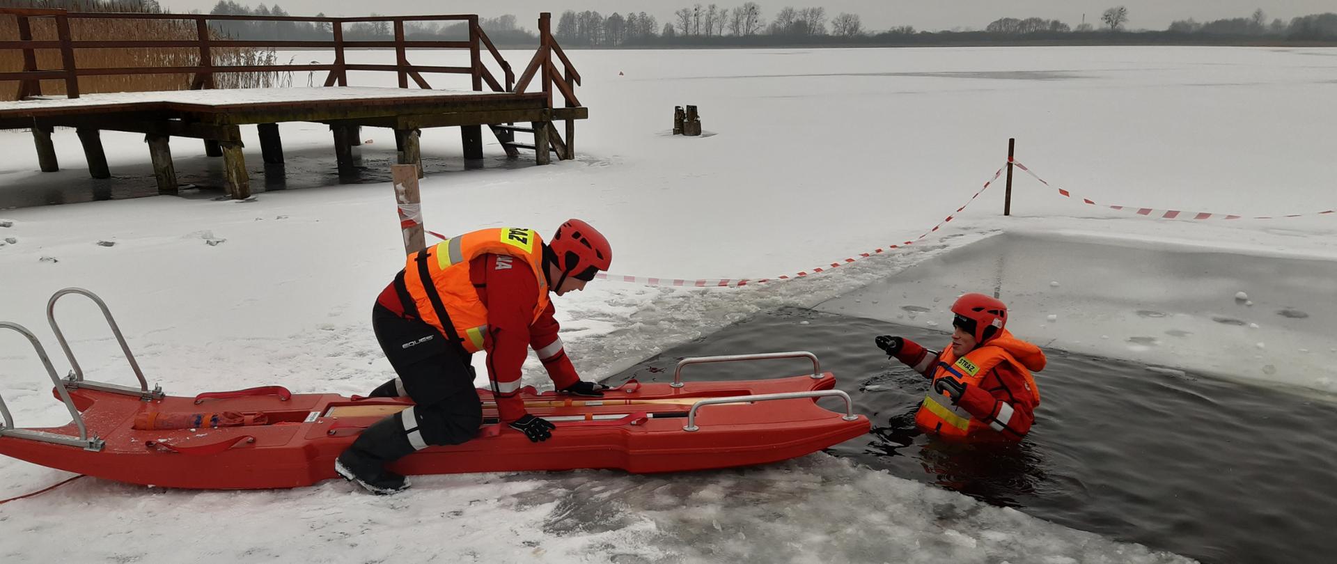 Zdjęcie przestawia dwóch ratowników na jeziorze w strojach niezatapialnych ćwiczących podejmowanie poszkodowanego z wody przy pomocy deski lodowej.
