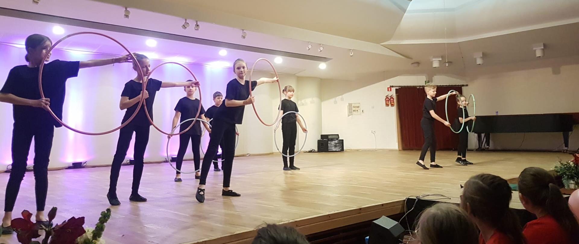 Ósemka dzieci ubranych na czarno tańczy trzymając przed sobą lub z wychyleniem w bok obręcze w kształcie koła.