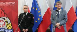 Mężczyzna w strażackim mundurze generalskim oraz drugi w okularach i szarym garniturze stojący przed mikrofonami. Za nim flagi Polski oraz Unii Europejskiej oraz logo Państwowej Straży Pożarnej