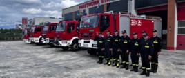 Strażacy stoją w szeregu obok stoją w szeregu samochody strażackie na tle komendy.