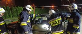 Na zdjęciu widać w porze nocnej 4 strażaków OSP, którzy zdają egzamin praktyczny z cięcia wraku samochodu przy pomocy narzędzi hydraulicznych. Strażacy ubrani są w umundurowanie specjalne, bojowe. W tle bloki mieszkalne miasta Ostrowiec Św.