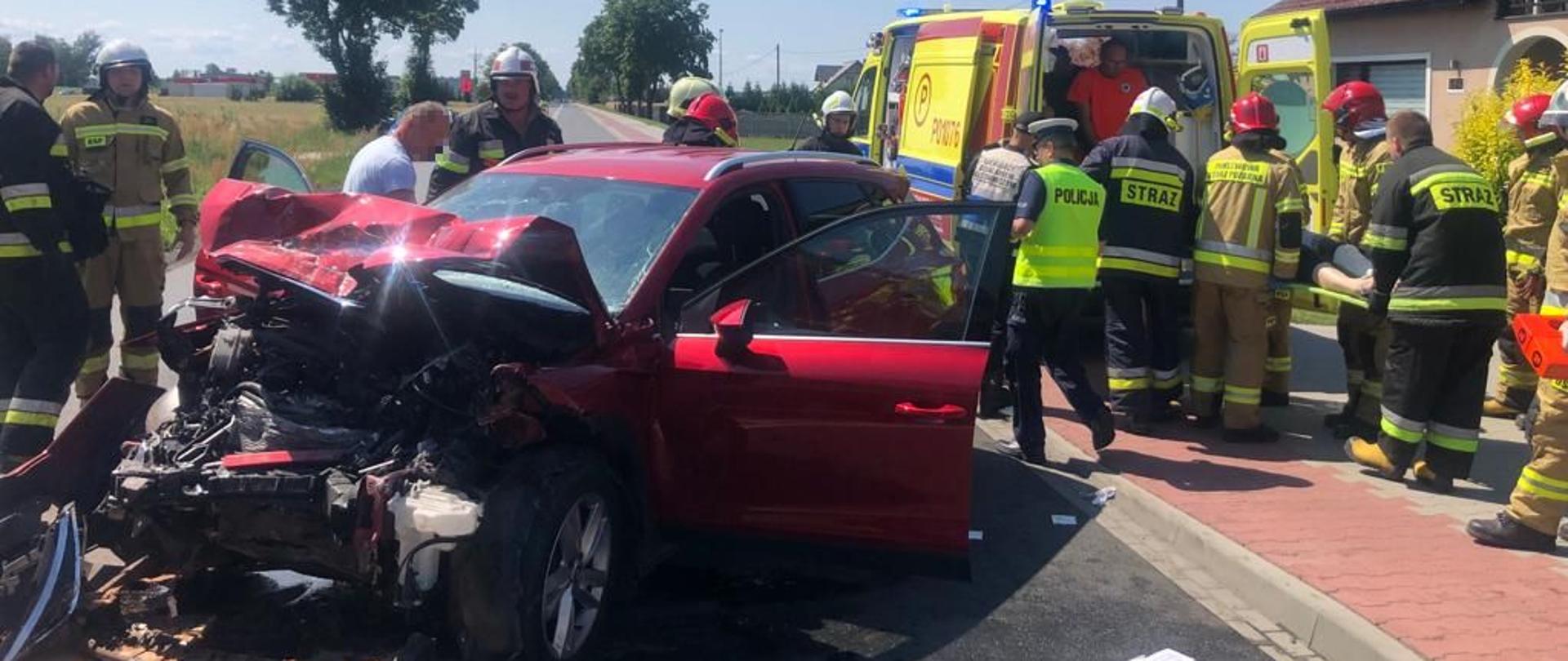 Seat z rozbitym przodem, obok policjant i strażacy niosący poszkodowaną na desce do karetki ZRM.