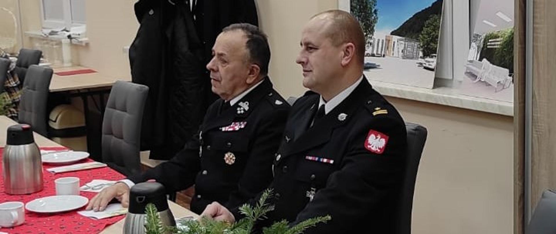 Na zdjęciu widać komendanta powiatowego PSP mł. bryg. Emila Szmyda oraz komendanta powiatowego OSP druha Tadeusza Niedźwieckiego. Siedzą Oni przy stole na którym znajdują się ozdoby świąteczne, szklanki, talerze i termosy. 
