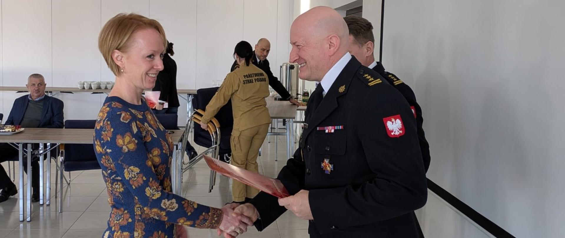 Z lewej strony kobieta, z prawej strażak w mundurze wyjściowym. Ściskają sobie dłonie, on podaje teczkę z dokumentami. Uśmiechają się. W tle widać krzątających się ludzi. 