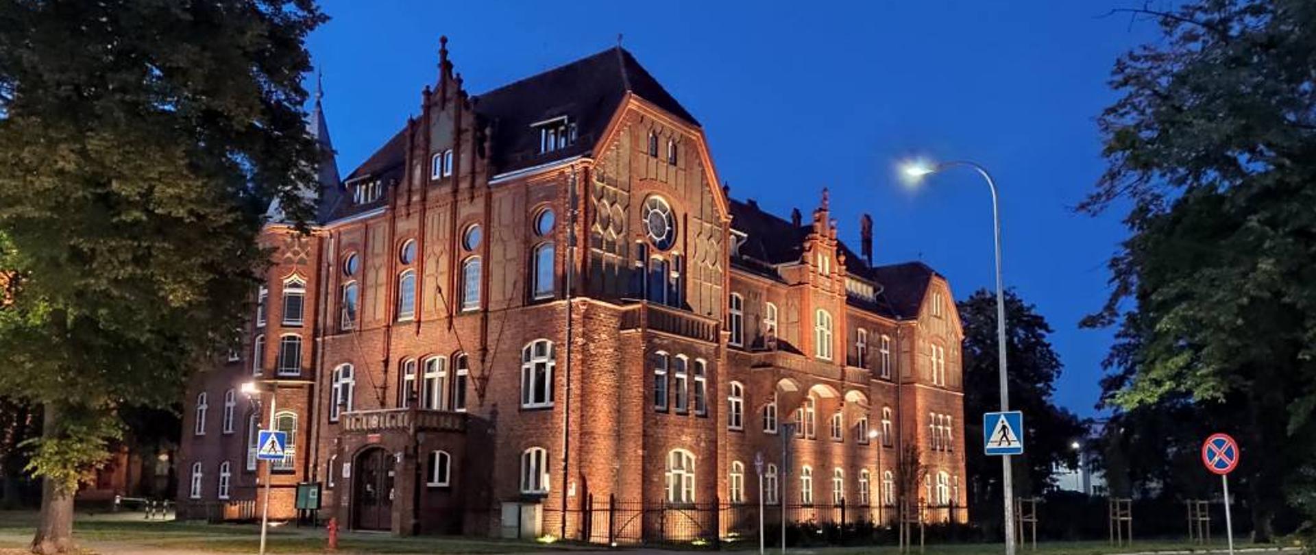 zdjęcie przedstawia od lewej strony oświetloną wieżę kościoła mariackiego w Szczecinku oraz oświetlony budynek szkoły muzycznnej na skrzyżowaniu ulic z czerwonej cegły nocą, na ciemno-niebieskim tle nieba. Po lewej i prawej stronie budynku zielone drzewa. Z prawej strony widoczne przejście dla pieszych