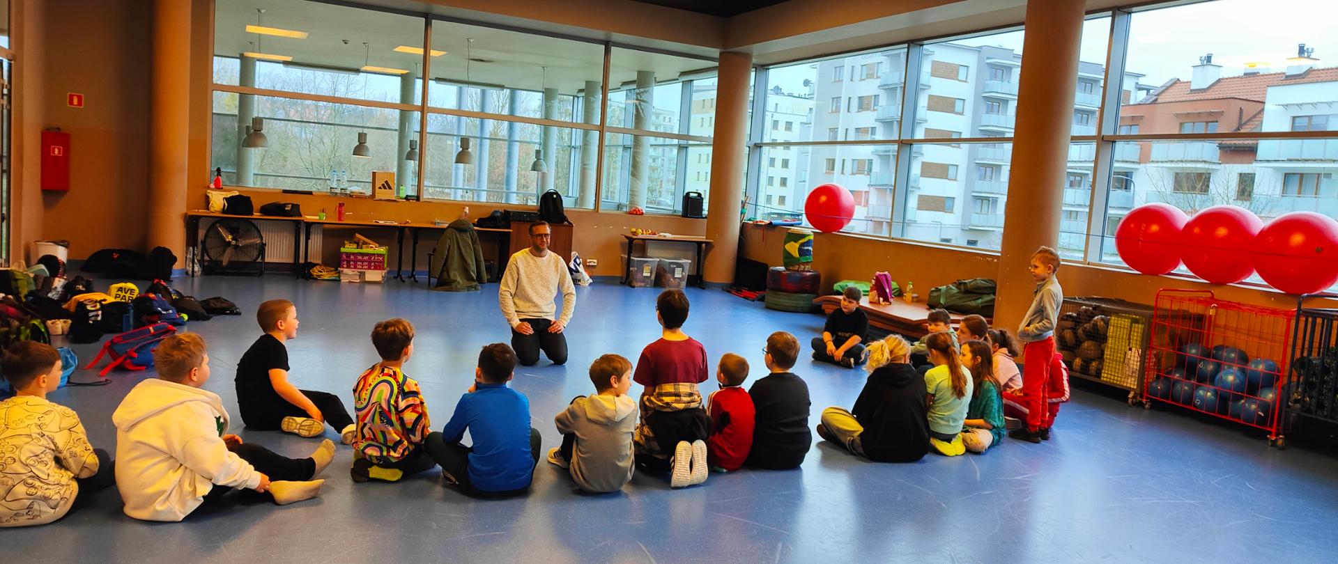 Spotkanie edukacyjne dla dzieci na półziomowisku w ramach akcji "Zdrowe ferie". Dzieci siedzą w kółku i słuchają edukatora.