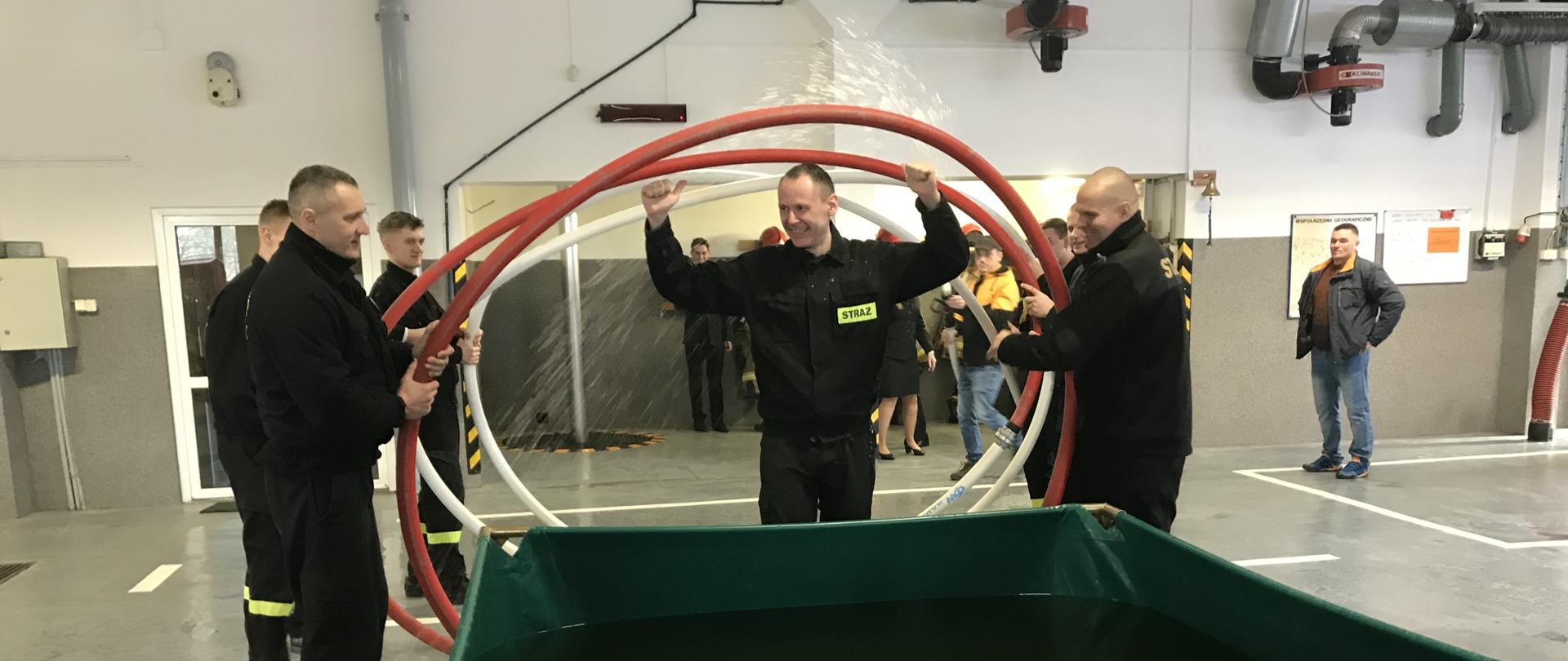 Zdjęcie zrobione na garażu Jednostki Ratowniczo-Gaśniczej w Kętrzynie. Strażacy ubrani w czarne ubranie koszarowe z zielono/żółtymi wstawkami odblaskowymi. Strażacy trzymają wąż strażacki zawinięty w spiralę przez którą przechodzi strażak z uniesionymi rękami. Idzie w kierunku zielonego zbiornika na wodę.