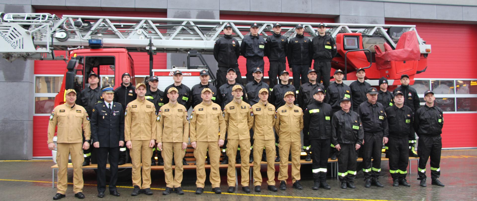 Zdjęcie przedstawia budynek Jednostki Ratowniczo-Gaśniczej Numer 1, przed którą stoi drabina mechaniczna stanowiąca tło dla strażaków. Wśród nich stoi 3 odchodzących funkcjonariuszy i Komendant Miejski PSP w Kielcach