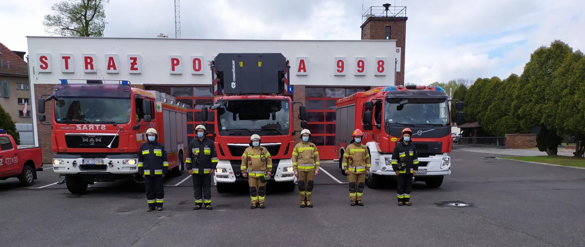 Strażacy stoją w rzędzie a w tle samochody i budynek