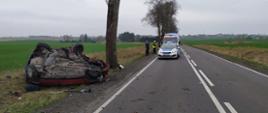 Zdjęcie przedstawia samochód osobowy po uderzeniu w drzewo oraz stojące w oddali na drodze samochody policji i Zespołu Ratownictwa Medycznego