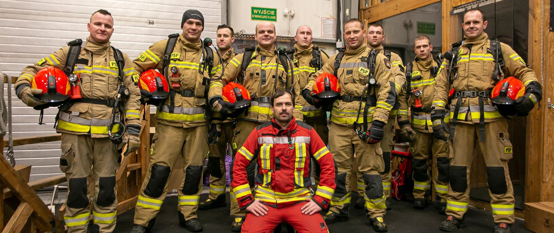 Na zdjęciu grupa strażaków w mundurach bojowych, hełmy czerwone trzymają w rekach. W raz z nimi na dole zdjęcia instruktor w czerwonym ubraniu bojowym.