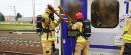 Peron dworca PKP w Rawiczu. Ze stojącego wagonu pasażerskiego wydobywa się dym. Obok niego na peronie znajdują się strażacy PSP i OSP prowadzący działania. Dwóch z nich przez wybite okno drzwi wagonu wkłada do jego wnętrza drabinę, na której przewieszona jest linia gaśnicza.Strażacy maja na sobie aparaty ochrony układu oddechowego. W tle torowisko i budynki.