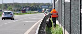 Zdjęcie przedstawia osobę ubraną w pomarańczową kamizelkę odblaskową, która porusza się wzdłuż pobocza autostrady, zbierając śmieci. Osoba ta trzyma duży czarny worek na śmieci i idzie obok metalowego ogrodzenia dźwiękochłonnego. Na drodze w tle widoczne są pojazdy, w tym srebrny samochód osobowy oraz czerwona ciężarówka. Po lewej stronie zdjęcia znajduje się pas zieleni oddzielający jezdnię od pobocza. W tle widoczne są drzewa oraz budynki, a dzień jest pogodny, z lekkim zachmurzeniem.