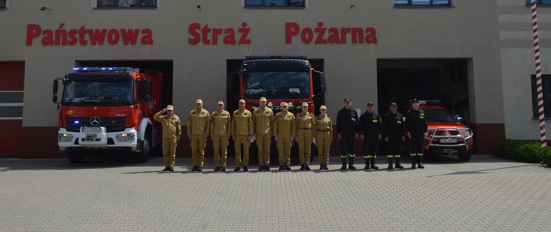 Plac przed komendą w Rawiczu. Na tle wozów bojowych, częściowo wystawionych przed bramami wyjazdowymi w szeregu stoja funkcjonariusze PSP. Stojący na prawym skrzydle zastępca komendanta oddaje honor poprzez salutowanie. 8 strażaków ma na sobie ubrania służbowe w kolorze piaskowym, pozostałych 4 - ciemne ubrania koszarowe. W pojazdach włączona jest sygnalizacja alarmowa.