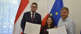 Przed białą ścianą stoją wiceminister Szeptycki, kobieta w czarnym ubraniu i mężczyzna w jasnoniebieskiej koszuli, trzymają otwarte teczki z dokumentami, za nimi pod ścianą flagi Polski, Austrii i UE, z lewej zasłonięte firanką okno.