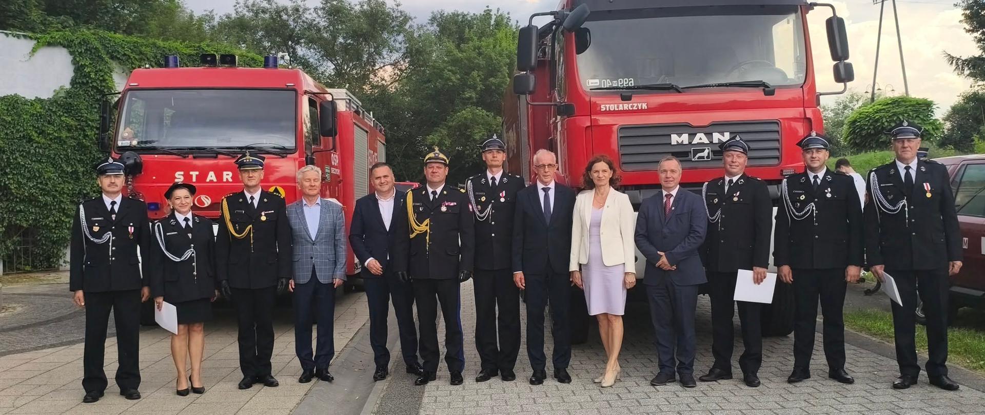 Pamiątkowe zdjęcie grupowe kilkunastu osób stojących w jednym rzędzie na wybrukowanym placu na tle dwóch dużych, czerwonych wozów strażackich. Po lewej stronie zaparkowany jest samochód marki Star, a po prawej większy wóz marki MAN z wyraźnym napisem „OSP RYBNO” nad przednią szybą. Wśród pozujących osób znajdują się strażacy i oficerowie w ciemnych mundurach galowych (w tym jedna kobieta w spódnicy mundurowej) oraz cywile – elegancko ubrani mężczyźni w garniturach i kobieta w jasnym komplecie ze spódnicą. Nad zgromadzonymi rozciąga się częściowo zachmurzone niebo, a w tle widać zielone drzewa.