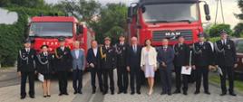 Pamiątkowe zdjęcie grupowe kilkunastu osób stojących w jednym rzędzie na wybrukowanym placu na tle dwóch dużych, czerwonych wozów strażackich. Po lewej stronie zaparkowany jest samochód marki Star, a po prawej większy wóz marki MAN z wyraźnym napisem „OSP RYBNO” nad przednią szybą. Wśród pozujących osób znajdują się strażacy i oficerowie w ciemnych mundurach galowych (w tym jedna kobieta w spódnicy mundurowej) oraz cywile – elegancko ubrani mężczyźni w garniturach i kobieta w jasnym komplecie ze spódnicą. Nad zgromadzonymi rozciąga się częściowo zachmurzone niebo, a w tle widać zielone drzewa.