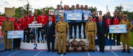 Szerokie ujęcie przedstawiające trzy zwycięskie drużyny XXIV Mistrzostw Województwa Podkarpackiego w Sporcie Pożarniczym stojące na podium rozstawionym na niebieskiej bieżni stadionu. Od lewej: KP PSP Stalowa Wola (miejsce 2), w centrum KM PSP Krosno (miejsce 1) z wielkim czekiem na 8 000 zł, od prawej KP PSP Brzozów (miejsce 3). Przed podium i obok niego stoją zaproszeni goście, samorządowcy oraz kadra dowódcza PSP w piaskowych mundurach służbowych.