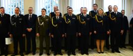 Na zdjęciu widzimy funkcjonariuszy stojących na zbiórce. Na zdjęciu przedstawieni Oficerowie ubrani są umundurowanie galowe którzy stoją w szyku podwójnym na tle okna. Kilku z nich trzyma w ręku okolicznościowe dyplomy i upominki. Wśród funkcjonariuszy stoi 1 osoba cywilna.