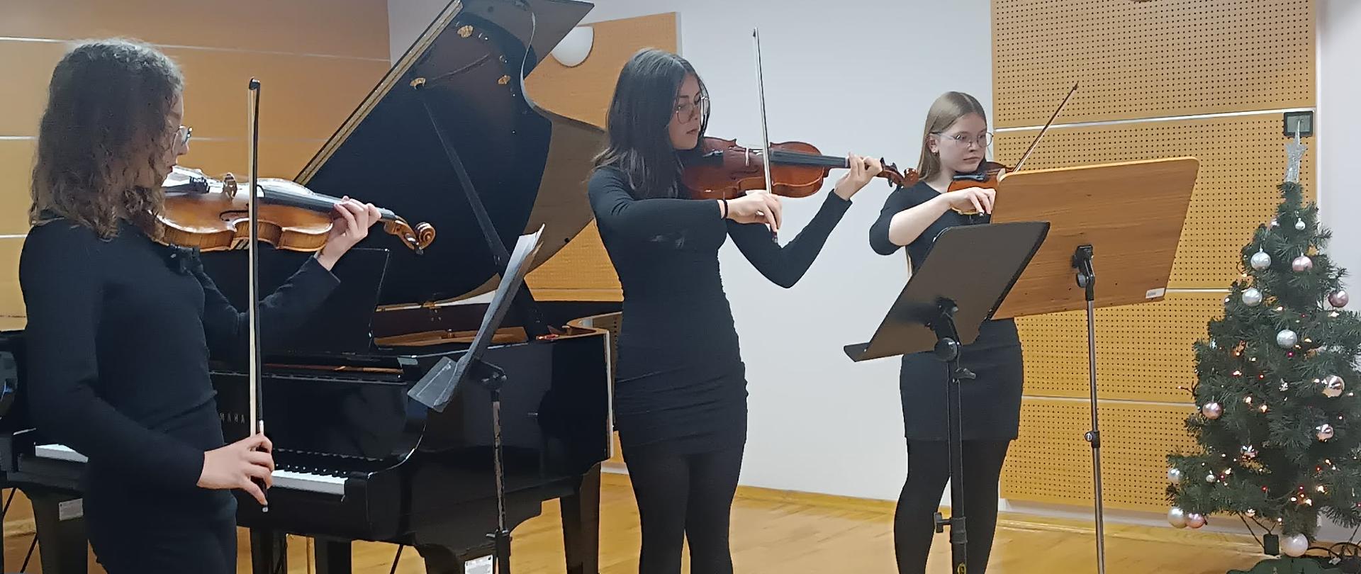 Zdjęcie przedstawia trzy uczennice grające na skrzypcach na scenie. Wszystkie trzy artystki są ubrane w eleganckie, czarne stroje. Stoją w różnych pozycjach, skupione na grze. Oprócz skrzypiec, po lewej stronie widoczny jest duży, czarny fortepian skrzydłowy. Przed skrzypaczkami ustawione są pulpity na nuty. Scena ma jasną, drewnianą podłogę. W tle, po prawej stronie, znajduje się udekorowana choinka, co sugeruje, że jest to koncert bożonarodzeniowy lub zimowy popis w szkole muzycznej. Zdjęcie oddaje moment artystycznego skupienia i profesjonalizmu podczas występu na żywo.