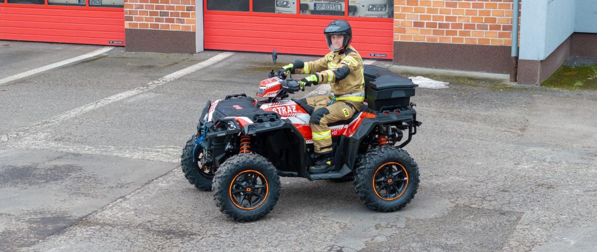 Strażak w pełnym umundurowaniu PSP siedzi na nowym quadzie ratowniczym Polaris Sportsman XP 1000 przed garażami jednostki, prezentując pojazd przeznaczony do działań w trudnym terenie.