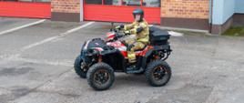 Strażak w pełnym umundurowaniu PSP siedzi na nowym quadzie ratowniczym Polaris Sportsman XP 1000 przed garażami jednostki, prezentując pojazd przeznaczony do działań w trudnym terenie.