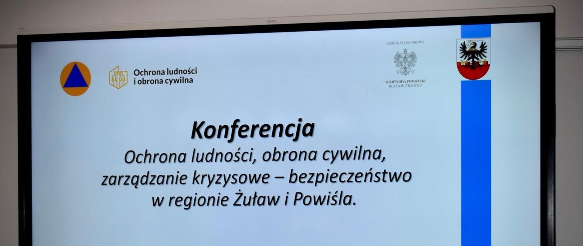 Slajd na ekranie konferencyjnym z napisem: Konferencja „Ochrona ludności, obrona cywilna, zarządzanie kryzysowe – bezpieczeństwo w regionie Żuław i Powiśla”